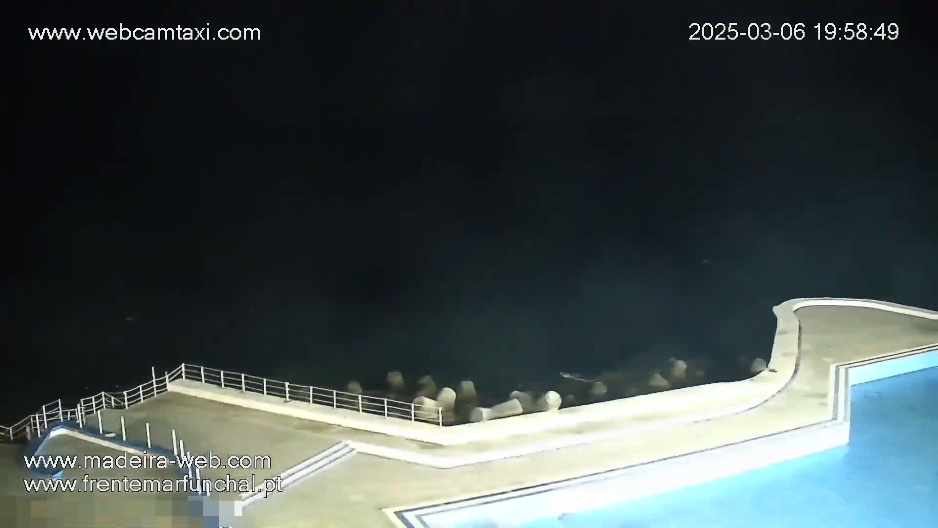 An outdoor nighttime view shows a pool deck with a section extending over dark water, where several large rocks are partially submerged.