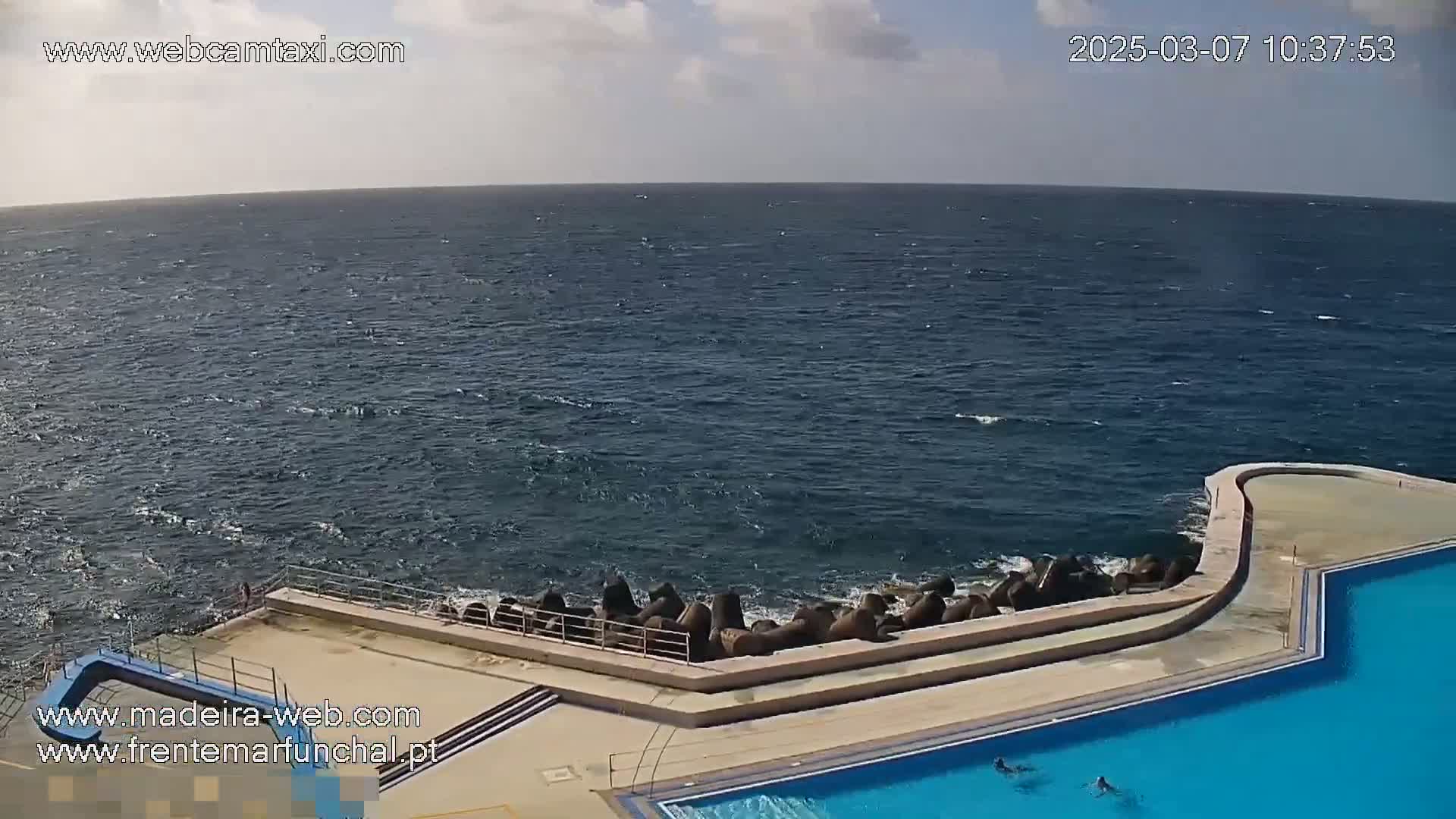 An outdoor swimming pool complex with a curved edge sits beside a rocky shore, where slightly choppy ocean water meets the land under a partly cloudy sky.