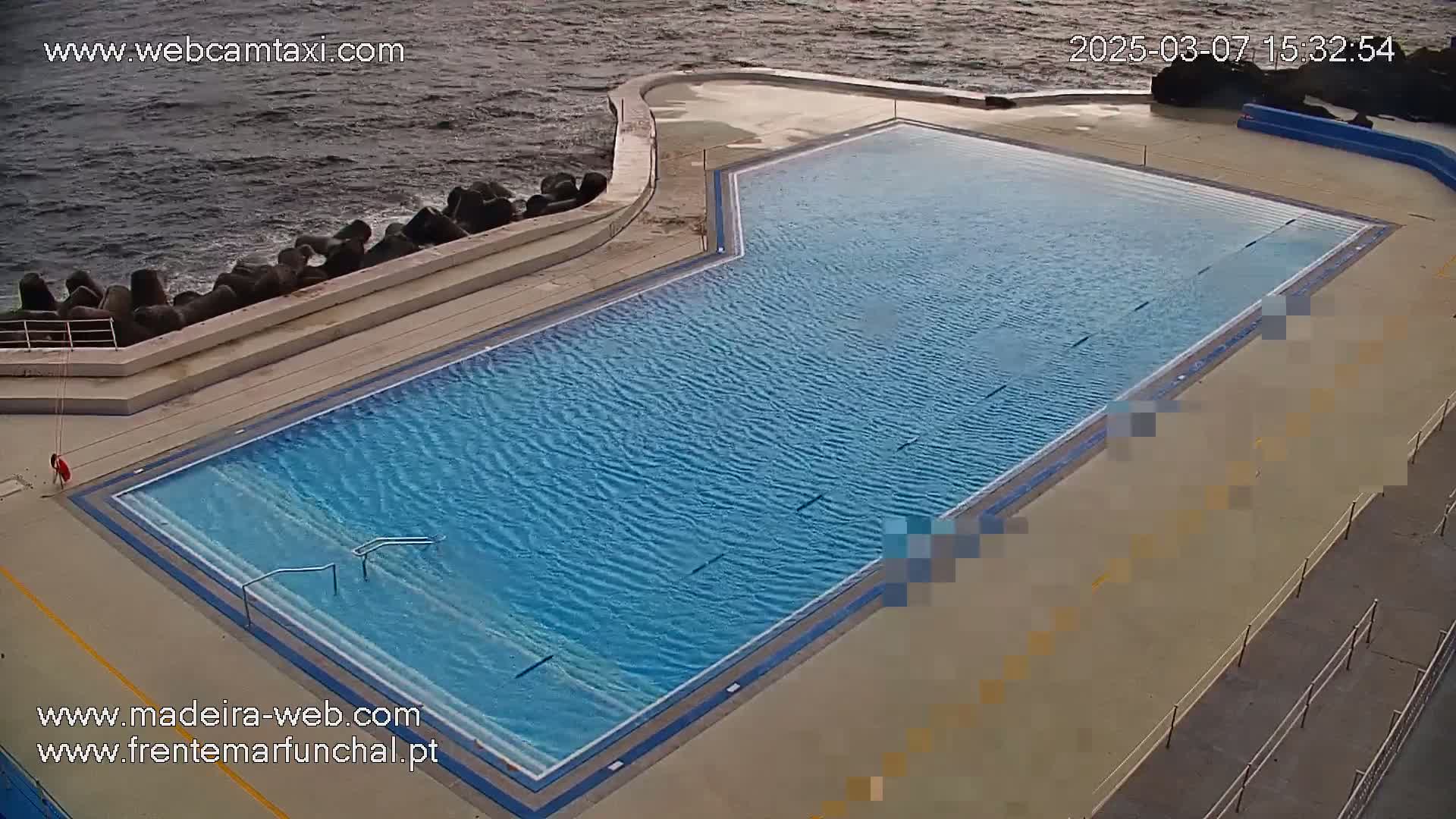 An outdoor pool with blue water sits next to a rocky coastline under partly cloudy skies.