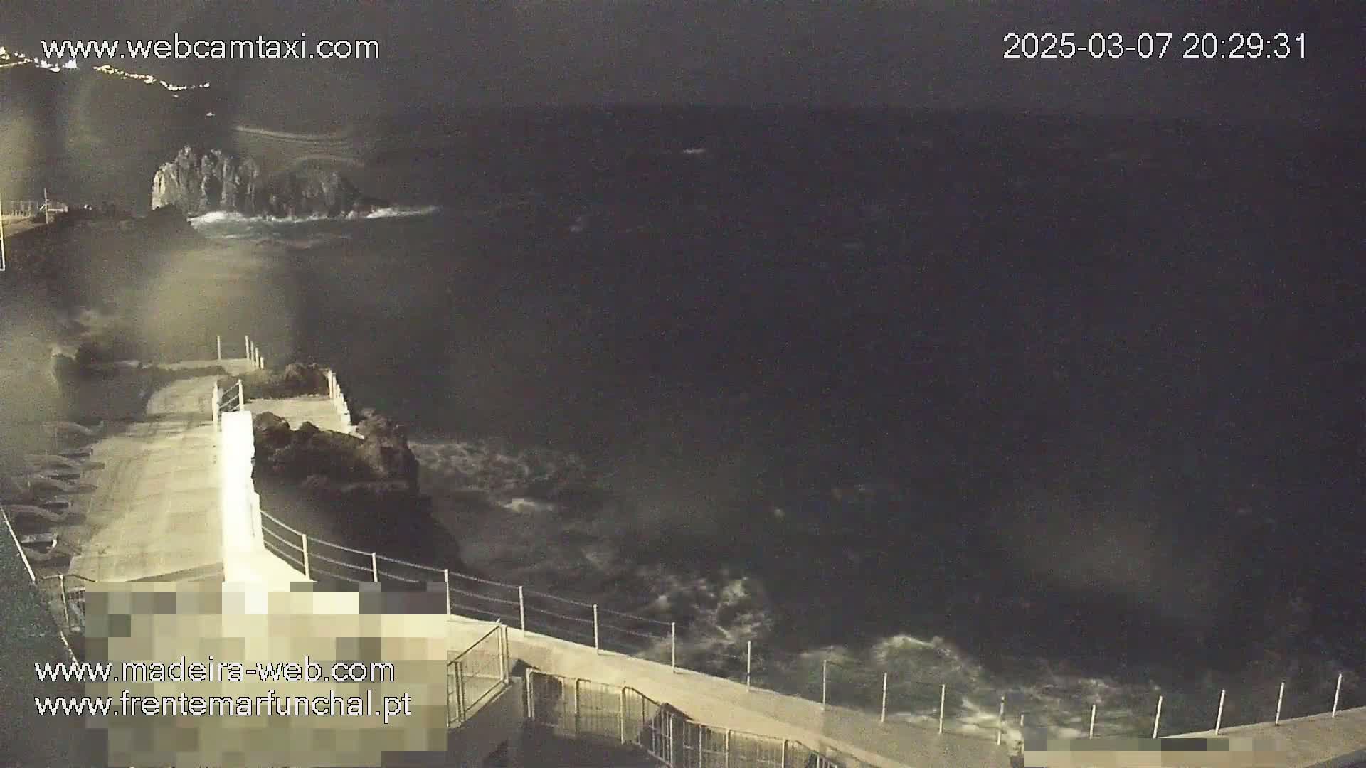 A nighttime view shows a rocky shoreline with a concrete walkway extending over the water, where dark waves crash against the rocks under a dark sky.