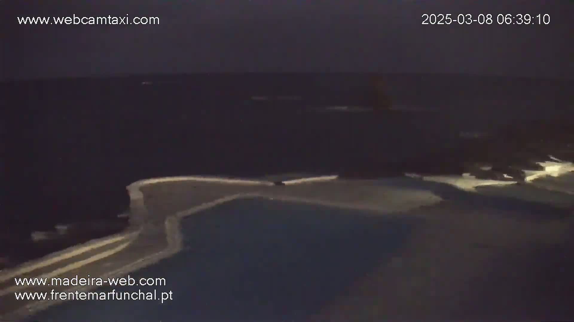A nighttime, dark view of a body of water and a paved area, possibly a dock or poolside, with faint light reflecting on the water.
