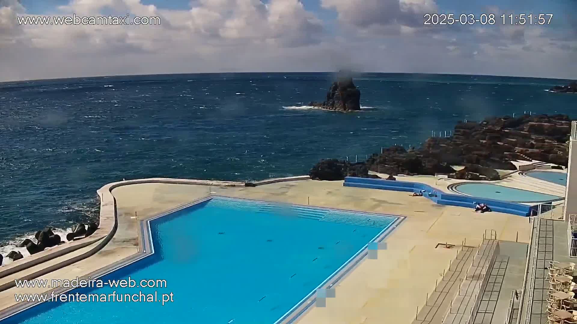 A large, blue, outdoor pool sits adjacent to a rocky coastline, overlooking a windy ocean.