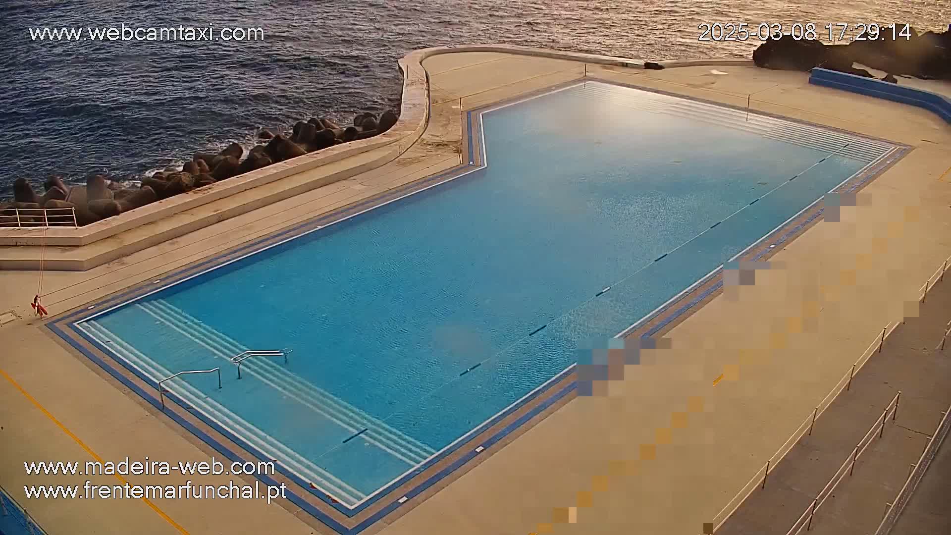An outdoor, rectangular swimming pool sits next to a rocky ocean shoreline under a mostly clear sky.