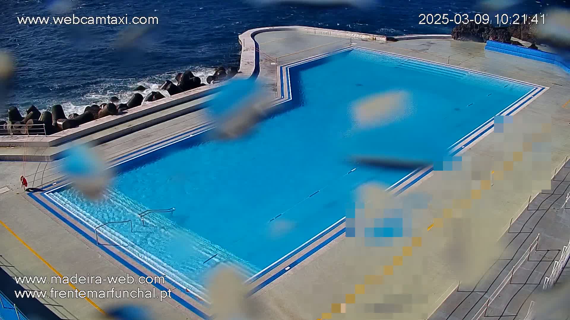 An outdoor oceanside swimming pool with a mostly clear blue surface is partially obscured by raindrops on a camera lens.
