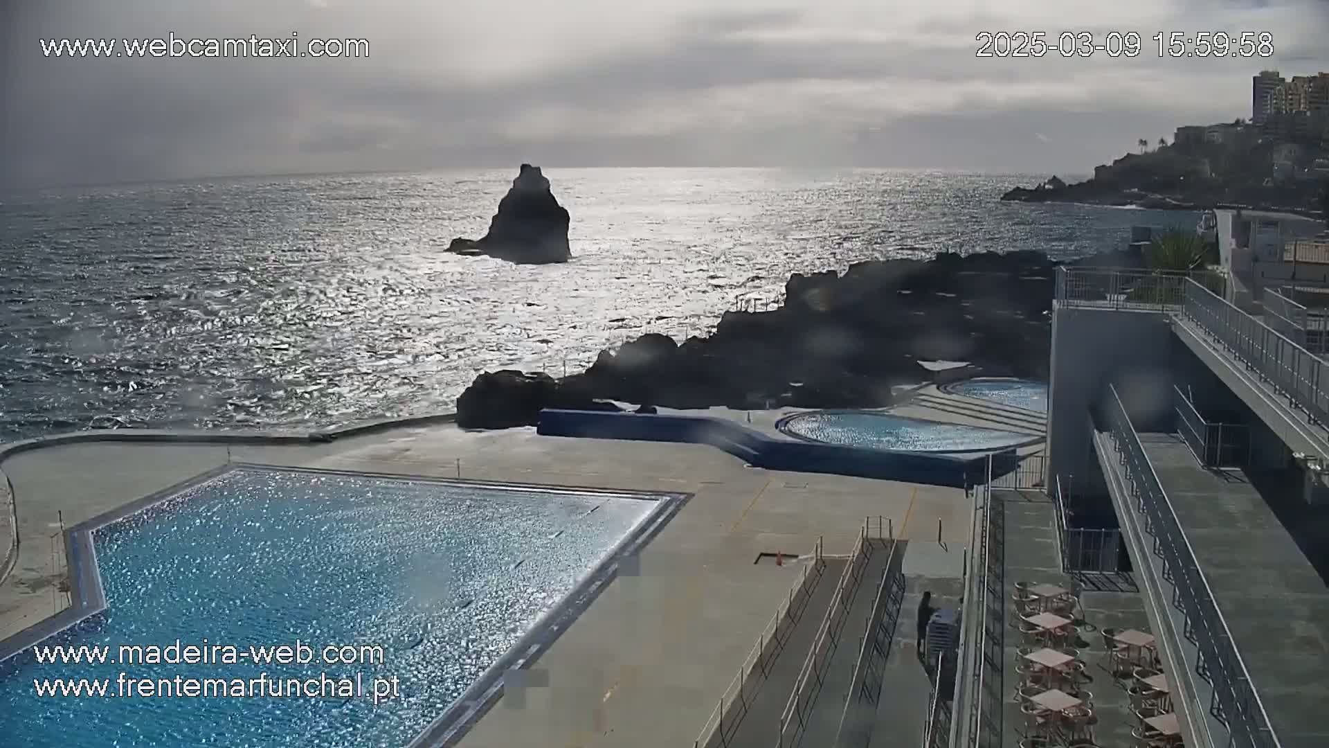 An outdoor pool complex overlooks a rocky coastline and choppy ocean under a cloudy sky.