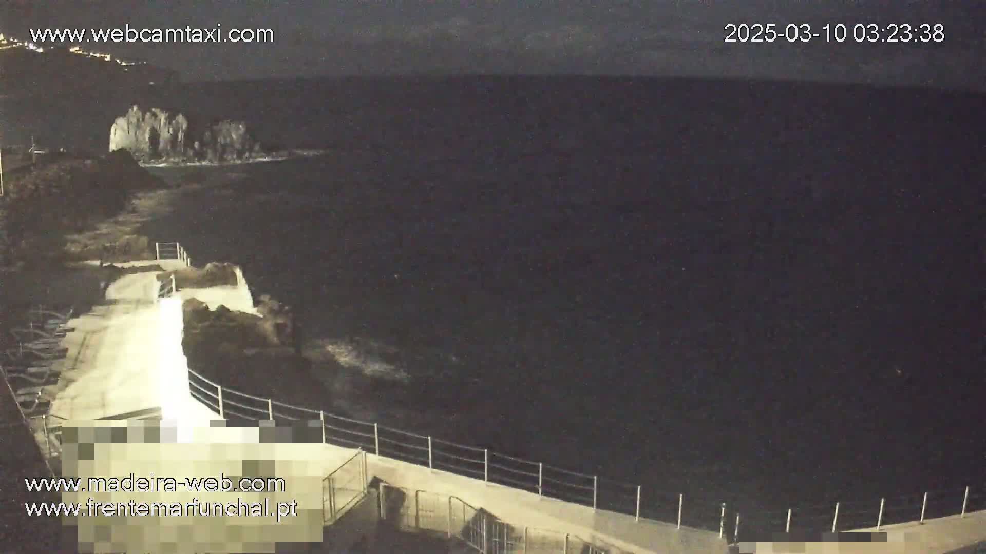 A nighttime coastal scene shows dark ocean waves lapping against a concrete walkway with a railing, near some rocky outcrops.