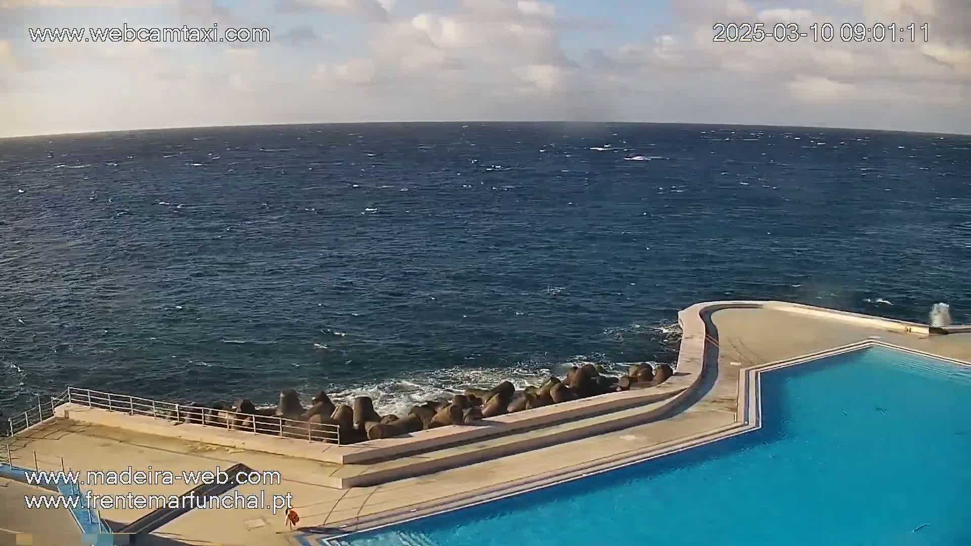 A partly cloudy sky overlooks a windy ocean with choppy waves crashing against a rock barrier next to an outdoor swimming pool.