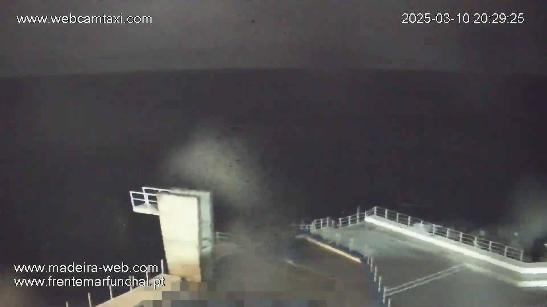 A nighttime, dark view of a waterfront structure with a railing, possibly a pier or dock, overlooking a dark body of water under a mostly clear, night sky.