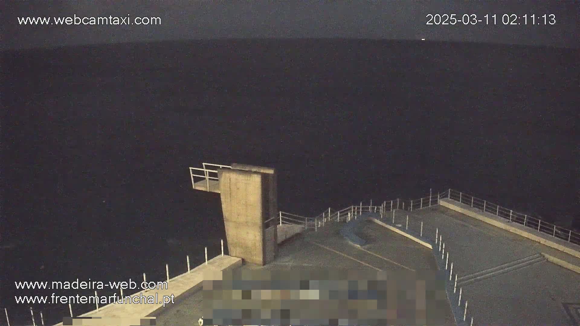 A nighttime, high-angle view shows a concrete diving platform extending over a dark body of water, with a railing surrounding a paved area.