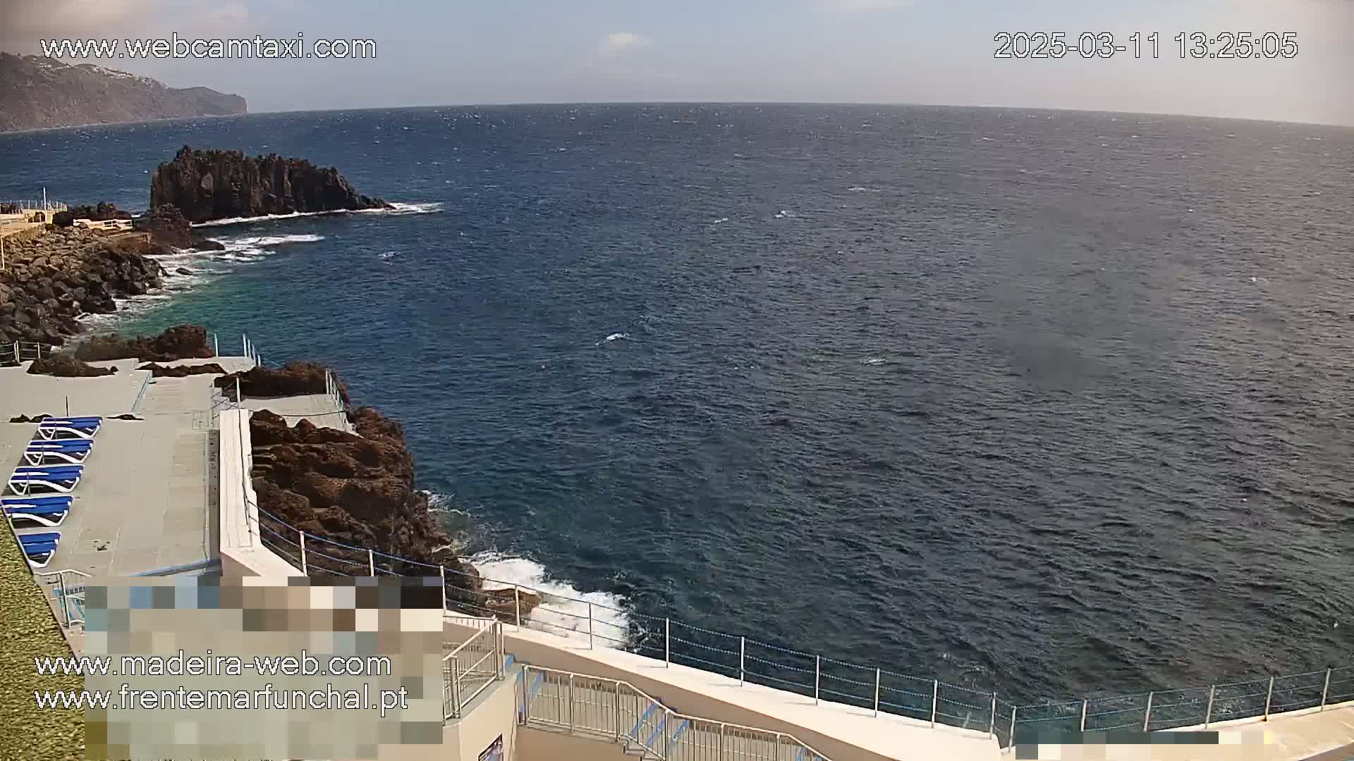 A mostly sunny day overlooking a rocky coastline with a small pool area and several lounge chairs, showing a dark blue ocean extending to the horizon.