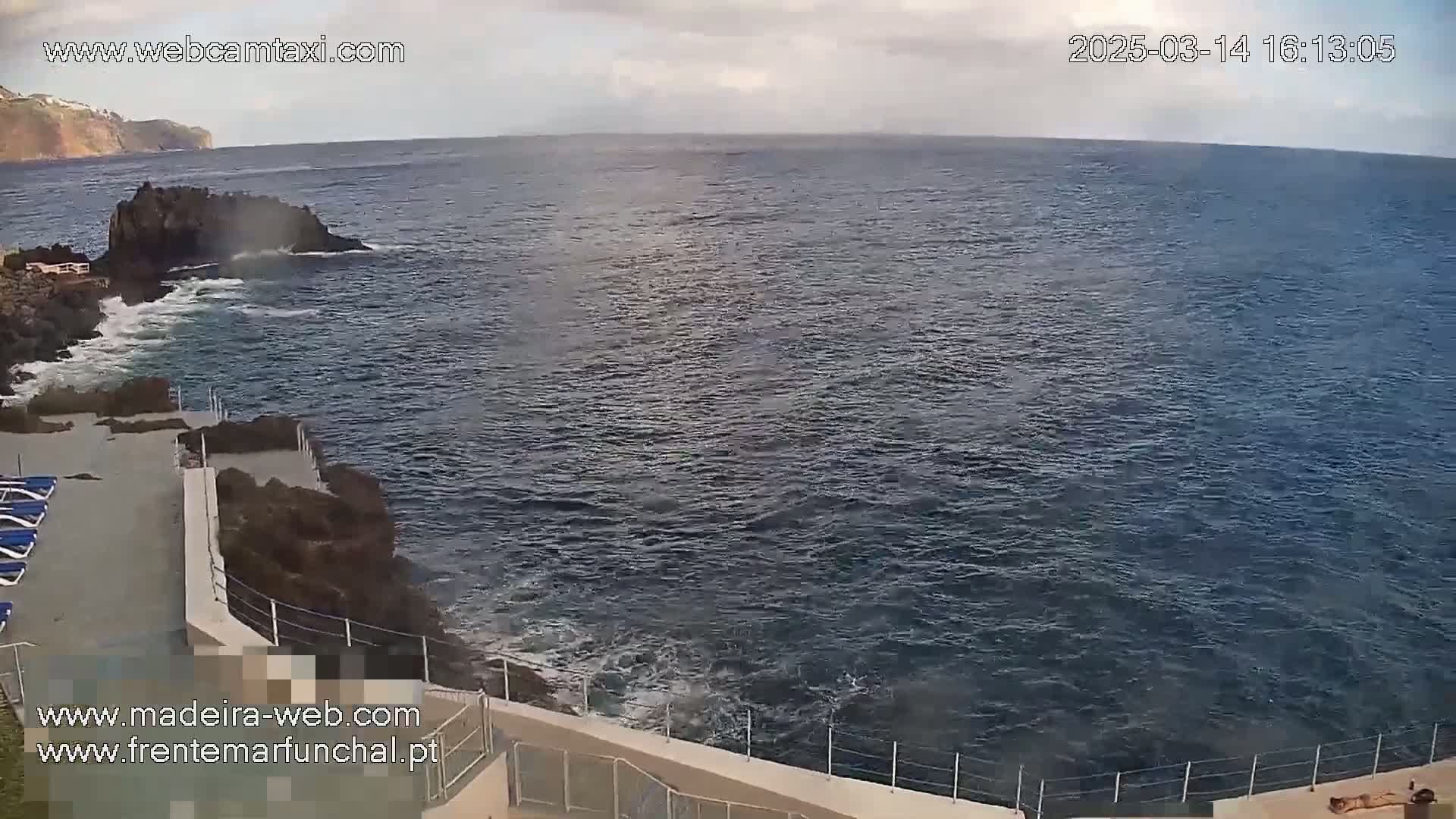 A rocky coastline with a concrete patio and lounge chairs overlooks a dark blue ocean with small waves under a partly cloudy sky.