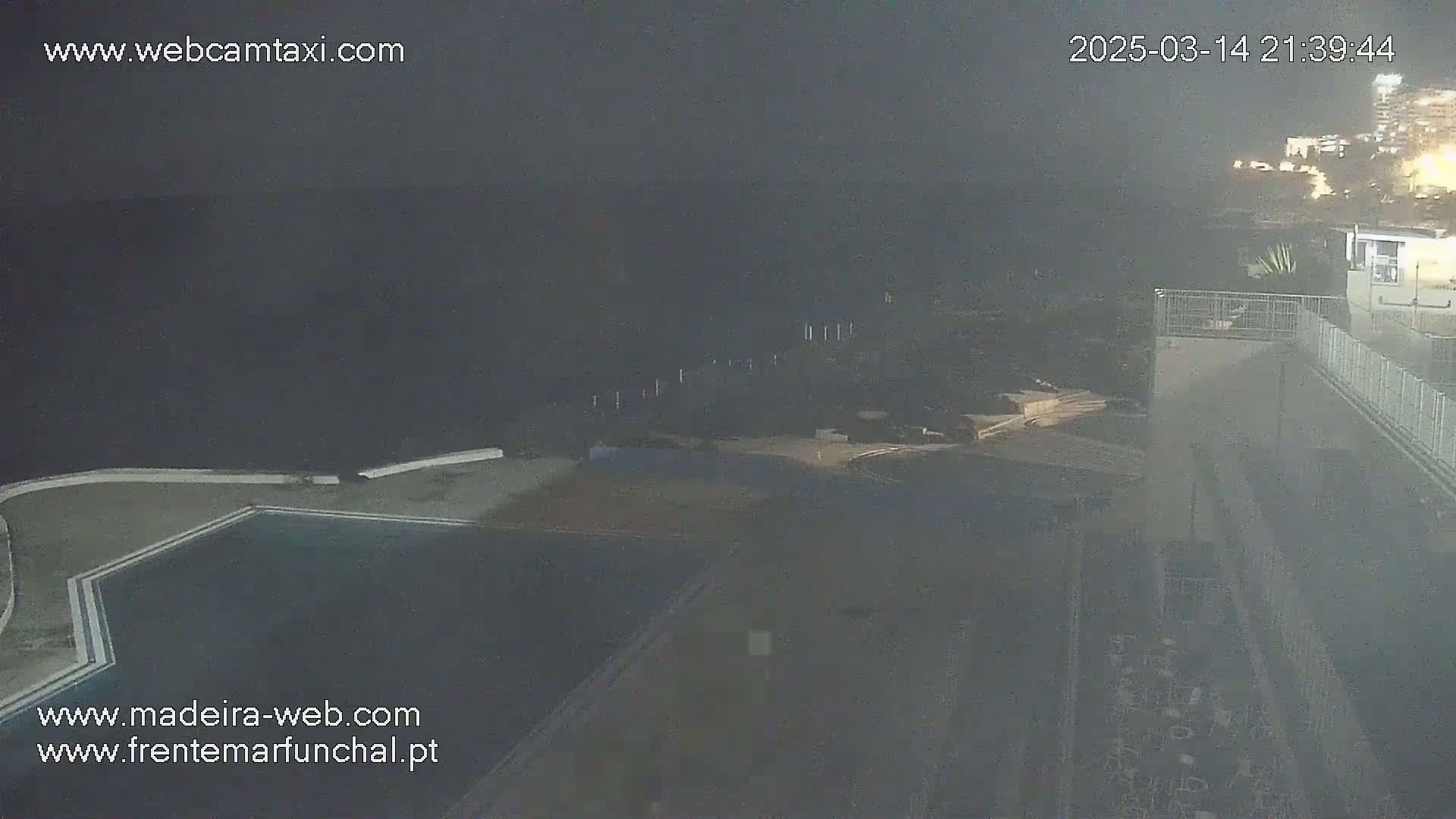A nighttime, hazy view shows an outdoor swimming pool and surrounding grounds near a coastline with buildings in the distance.