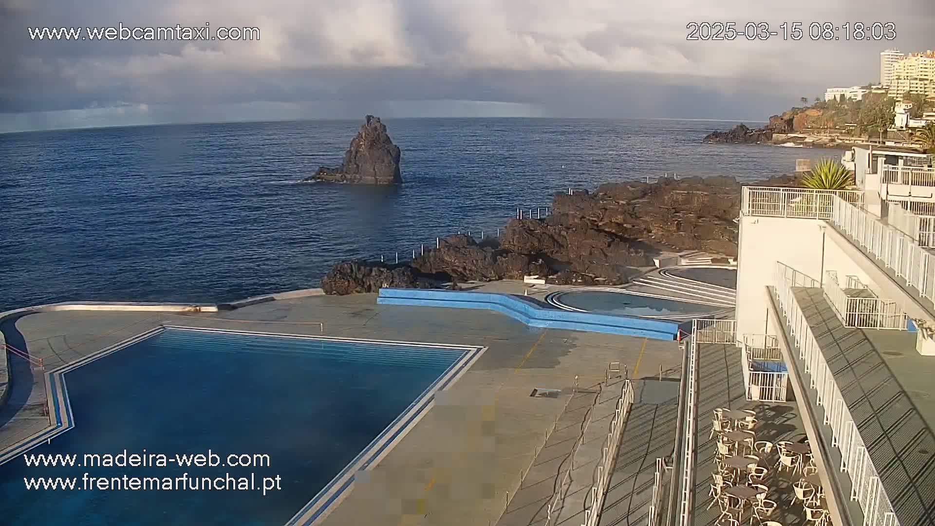 An outdoor scene shows a large swimming pool next to a building overlooking a rocky coastline and dark blue ocean under a partly cloudy sky.