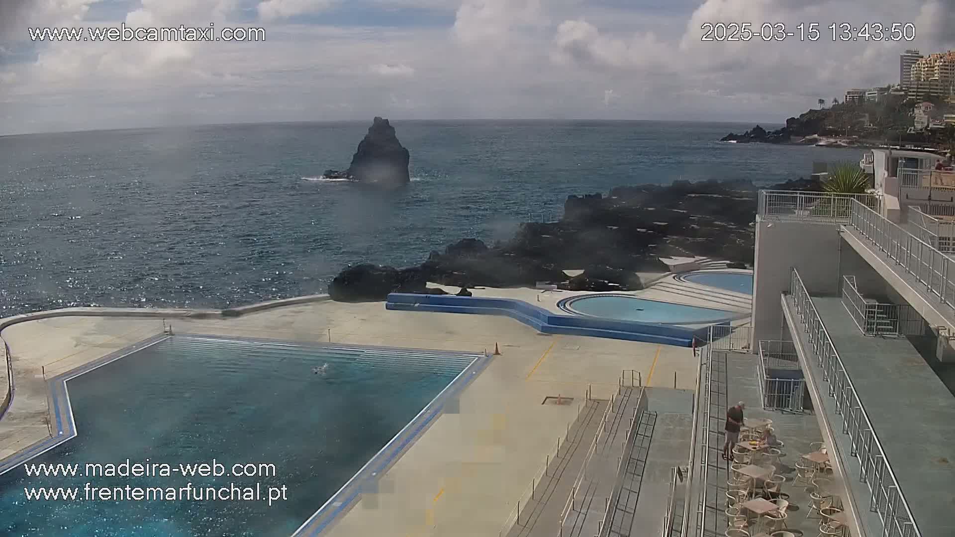 An outdoor pool complex overlooks a rocky coast and ocean under partly cloudy skies.
