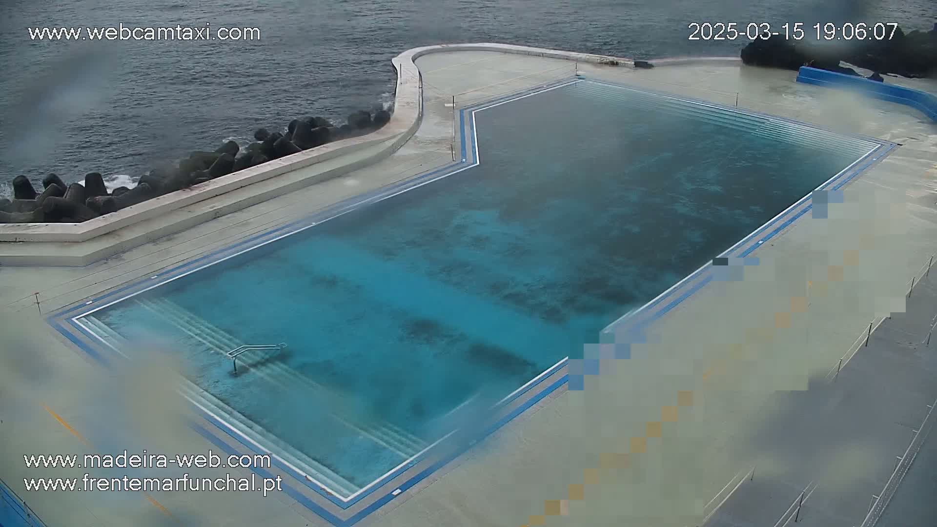 An outdoor seawater pool with steps leading into the water is situated next to a rocky ocean shore on an overcast day.