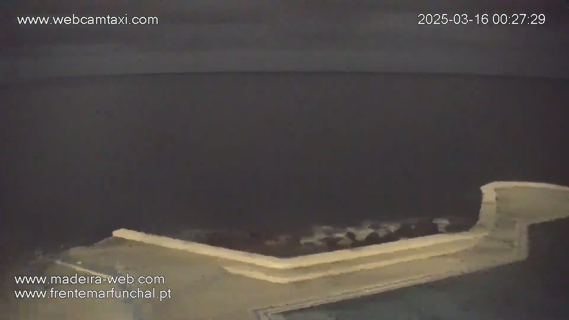 A low-light nighttime view shows a portion of a light-colored structure overlooking a dark body of water with small waves.