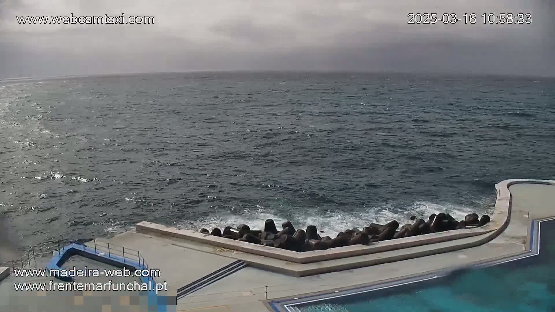 A mostly cloudy day shows a dark-blue ocean with small waves crashing against a concrete seawall next to a swimming pool.