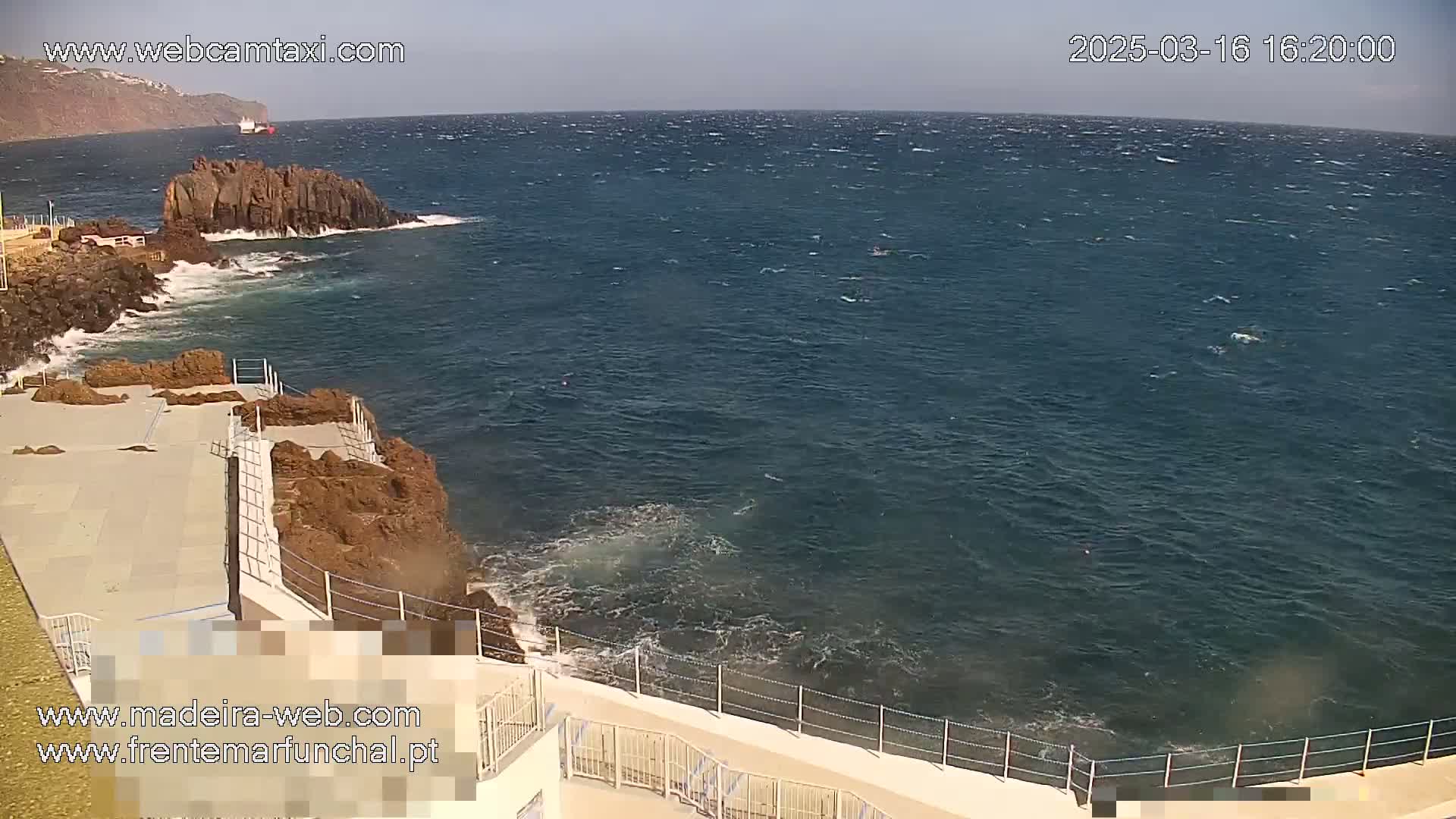 A rocky coast with a paved area and railings overlooks a dark blue, windy ocean with small waves crashing against the rocks.