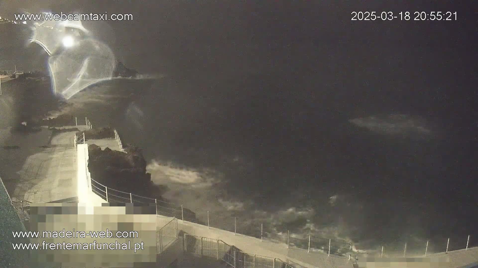 A nighttime, high-angle view shows dark ocean waves crashing against a concrete wall with a metal railing.