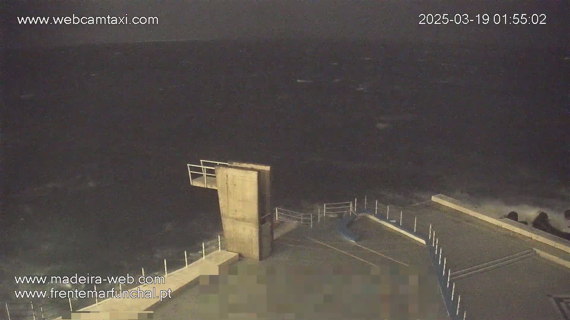 A nighttime view of a concrete diving platform overlooking a dark, choppy ocean under a cloudy sky.