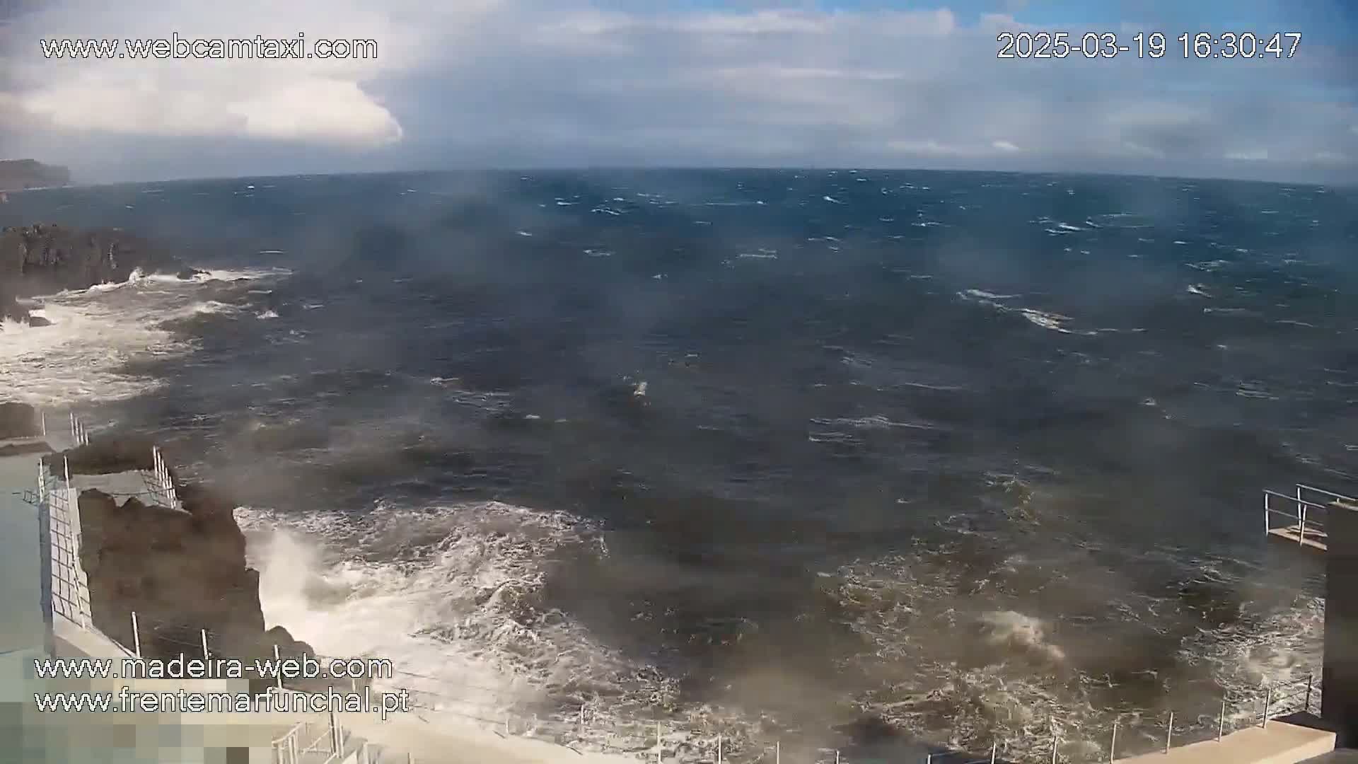 Rough, dark-colored waves crash against a rocky coastline on a partly cloudy day.