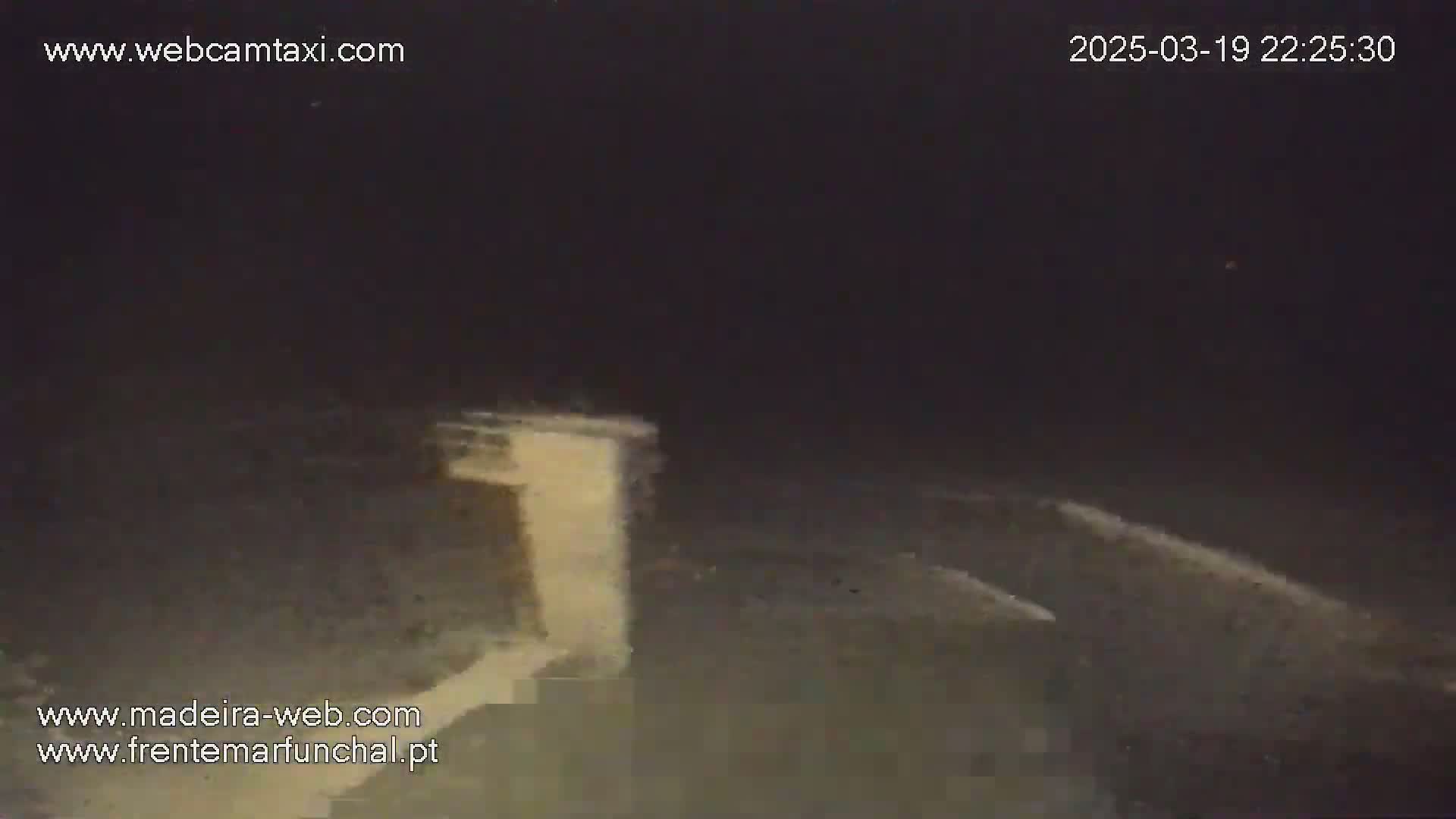 A nighttime, low-light view shows a light-colored structure, possibly a wall or building, extending from the bottom to the middle of the frame, with a dark, possibly paved, area extending from the middle to the bottom of the image.