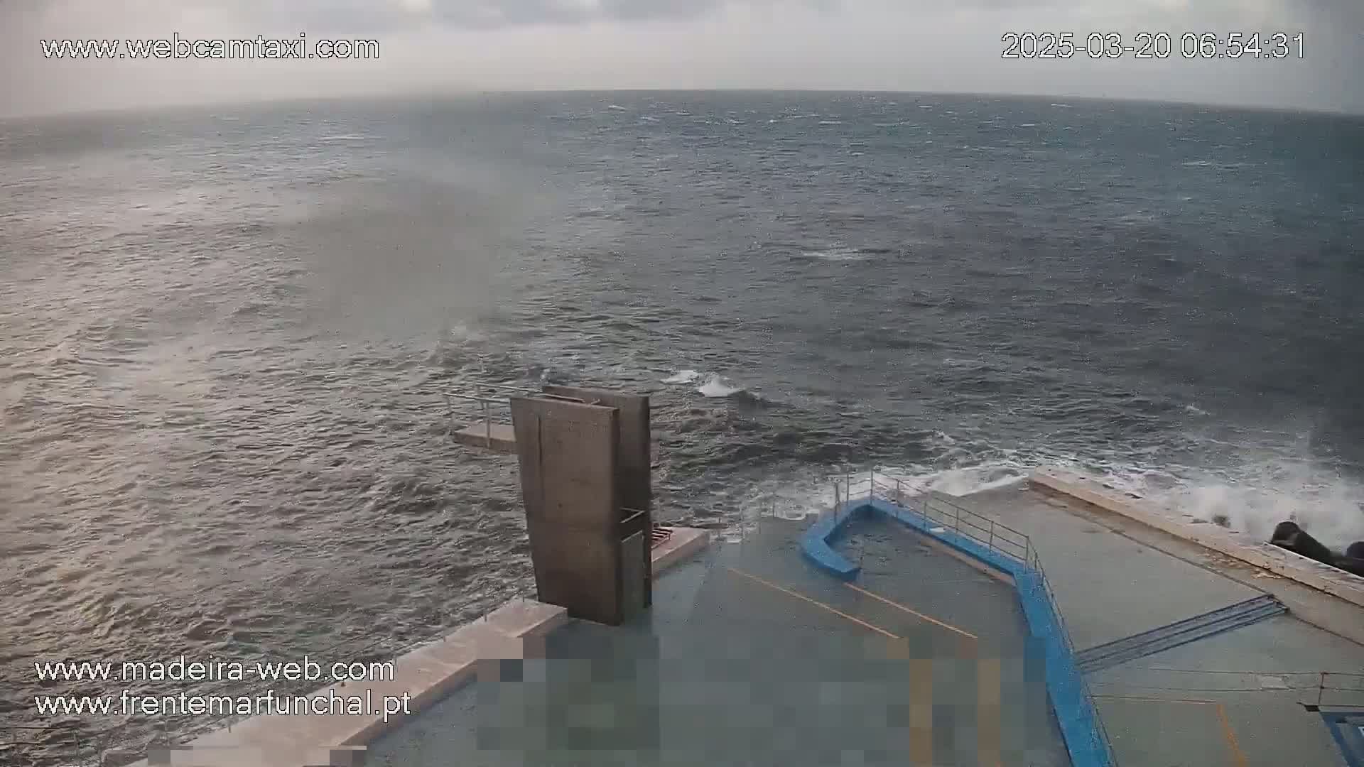 A concrete diving platform and adjacent area are partially submerged by choppy, gray water under an overcast sky.