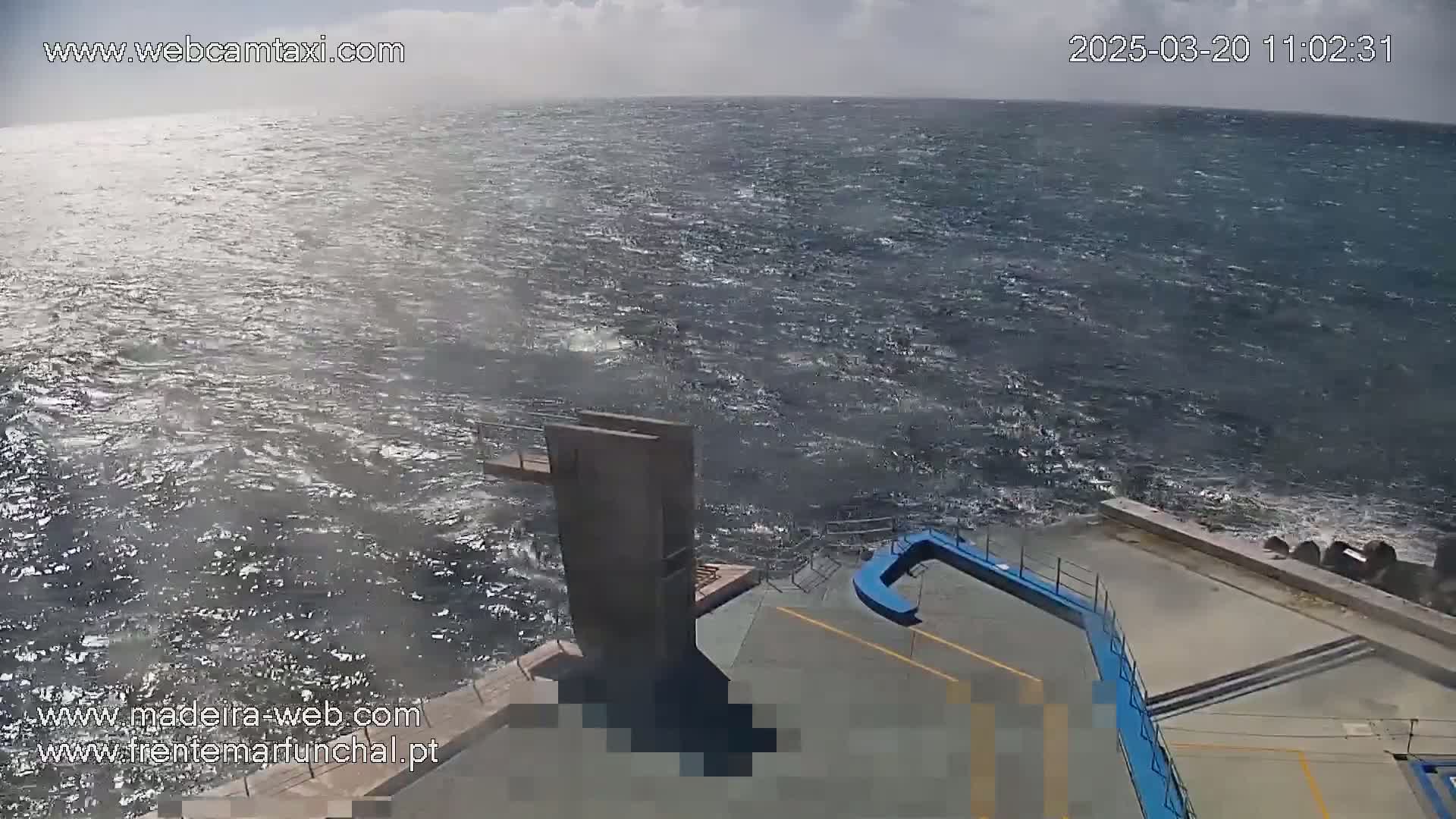 A windy day shows choppy ocean waves crashing against a concrete platform with a diving board and other structures.