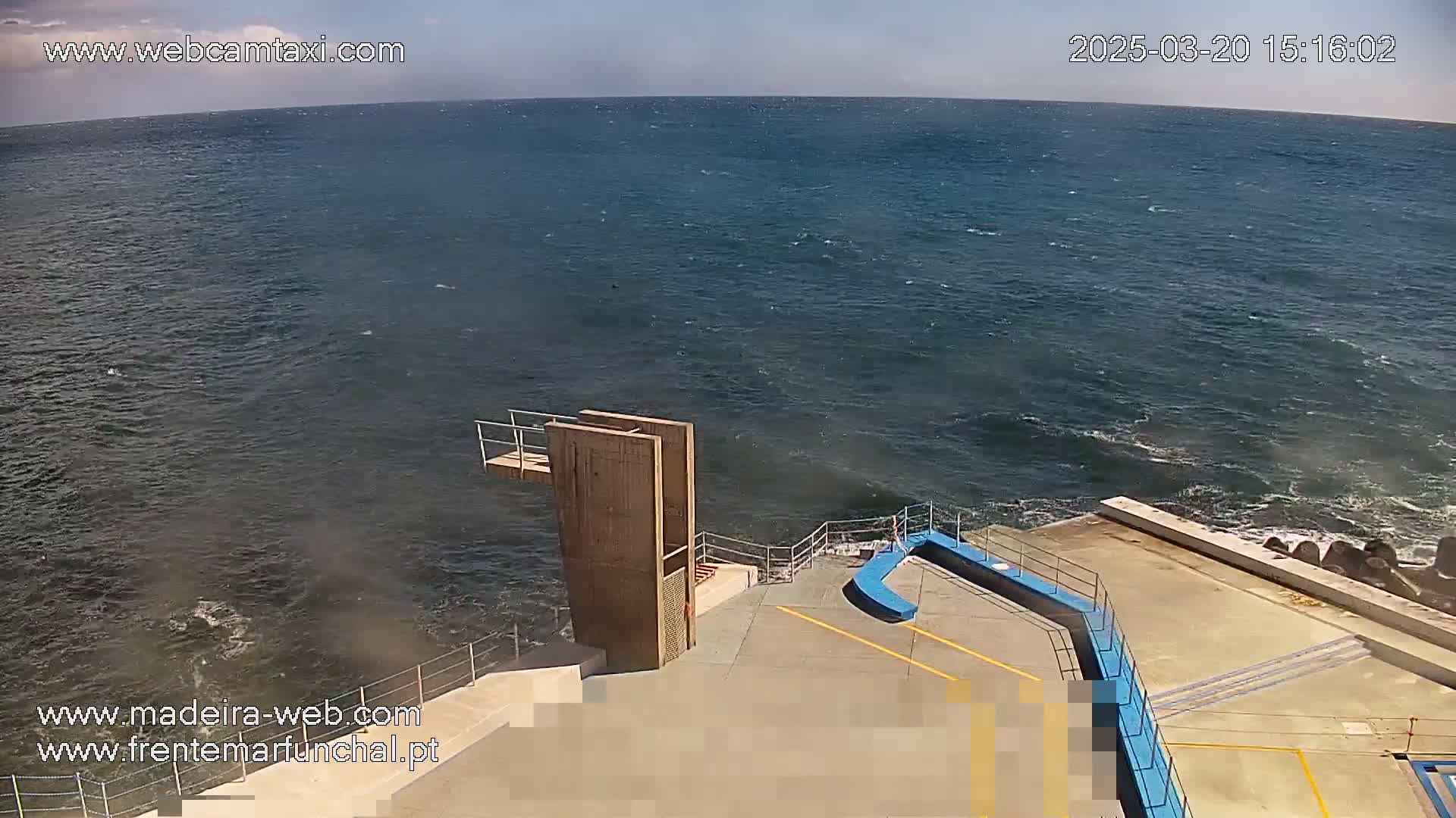 A wooden diving platform sits on a concrete area next to a choppy blue ocean under a mostly clear sky.