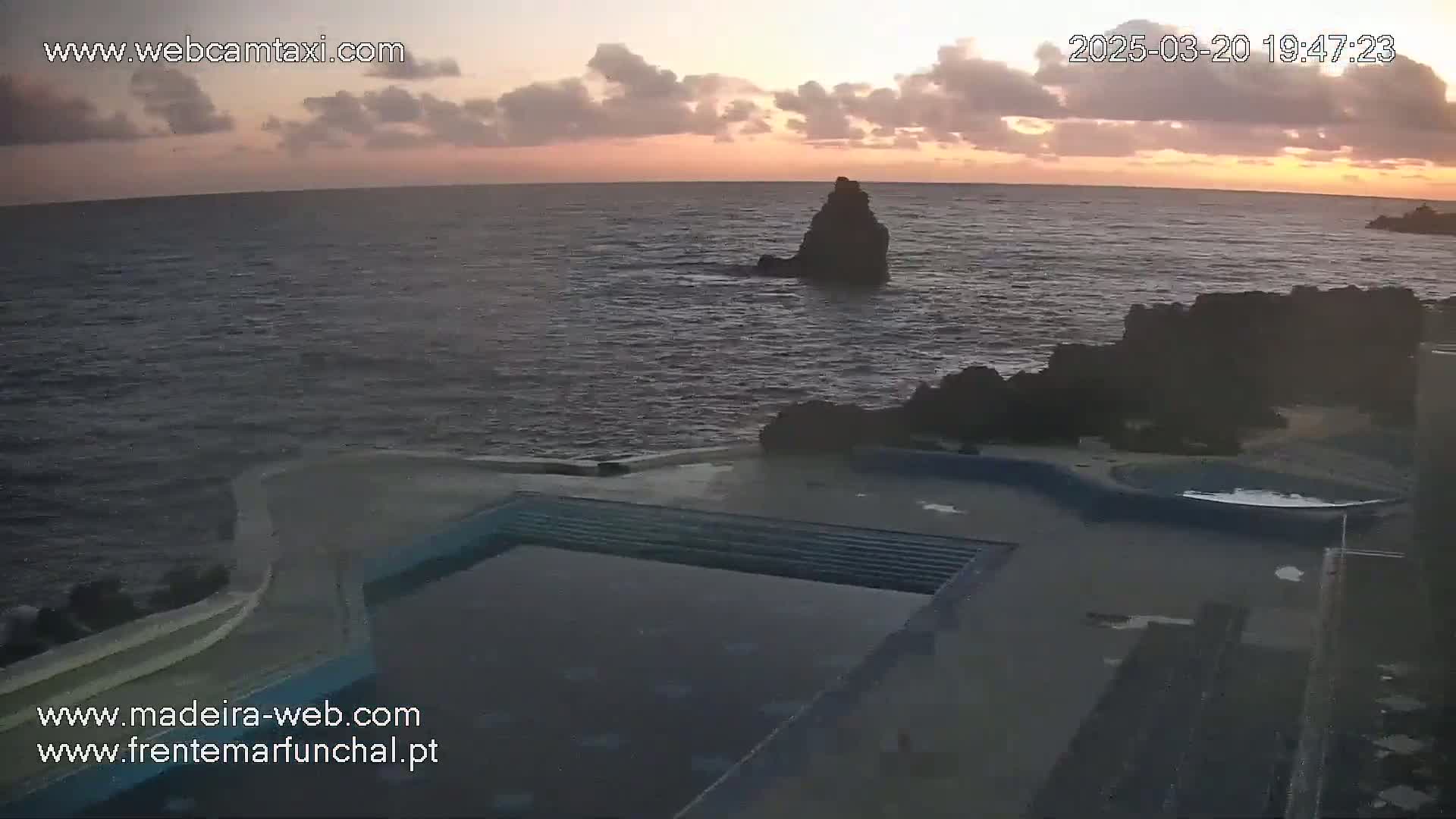 An outdoor pool sits near the ocean under a partly cloudy sunset sky.
