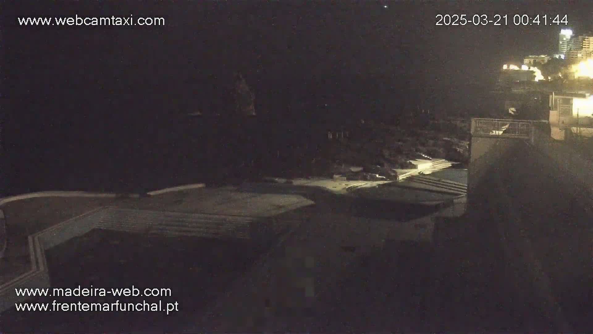 A nighttime, high-angle view shows a dark, rocky coastline with buildings and some lights visible in the distance.