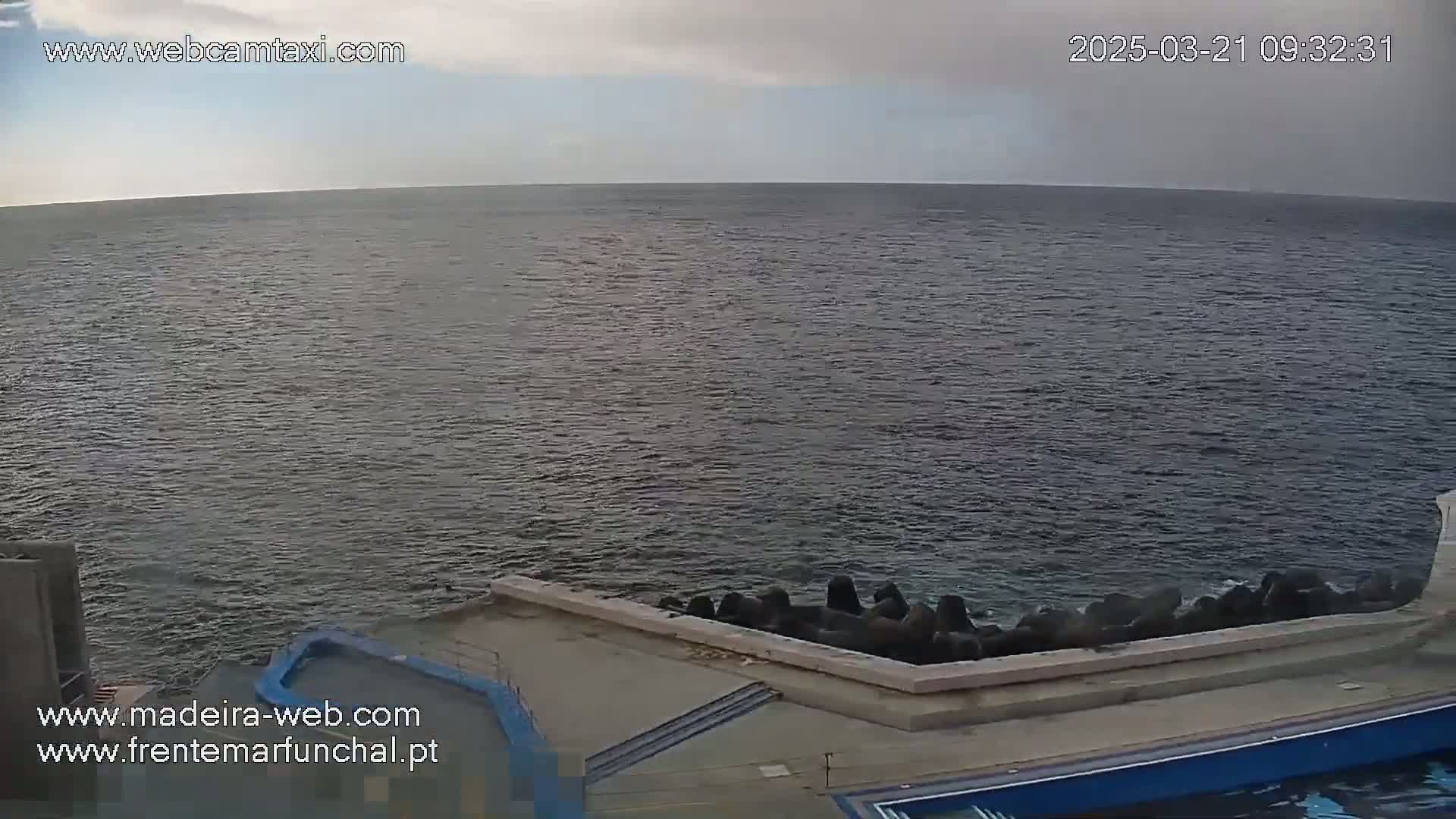 A mostly cloudy day shows a calm ocean meeting a low, concrete seawall with dark rocks at the water's edge.