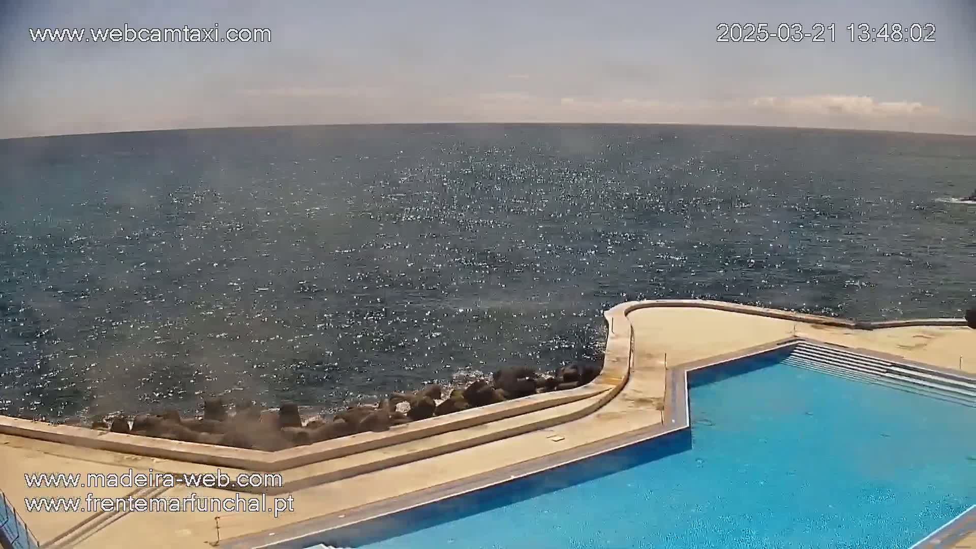 A mostly sunny day shows a wavy ocean next to an outdoor pool with stone steps.