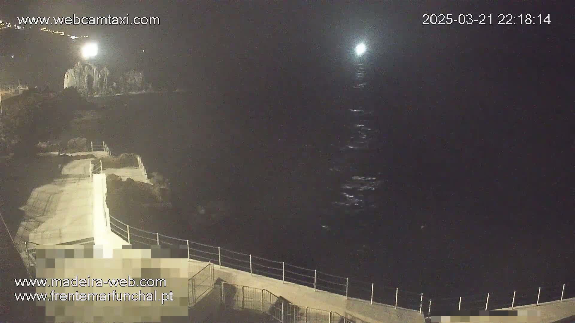 A nighttime view of a concrete walkway and railing overlooking dark ocean water under a partly cloudy sky.