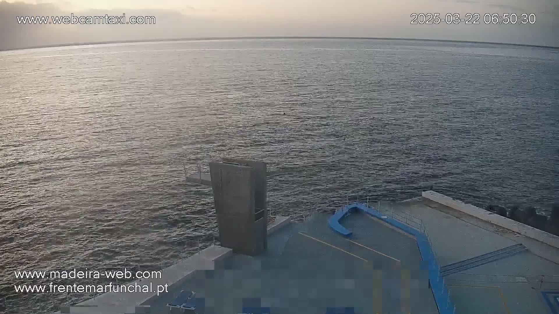 A calm body of water is seen from an elevated vantage point, showing a diving platform and surrounding paved area at dawn or dusk.