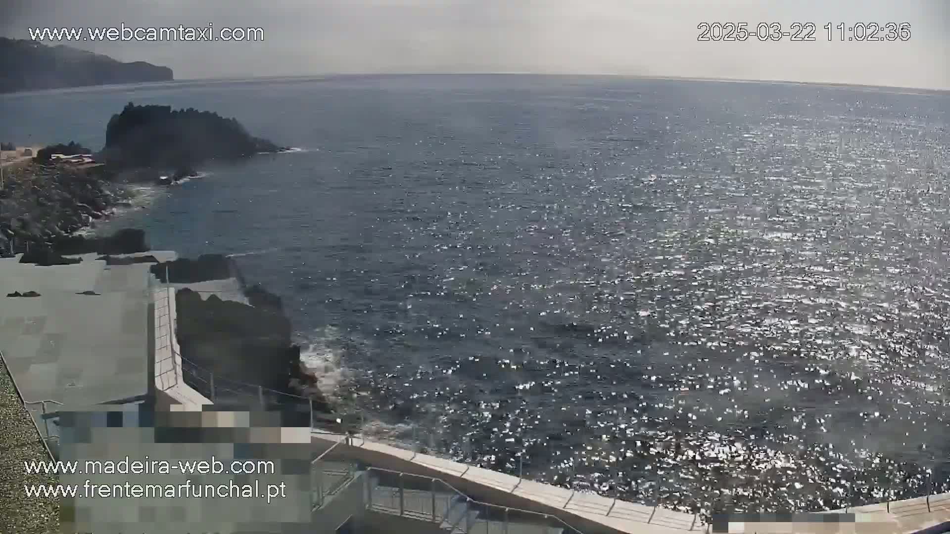 A rocky coastline with a paved walkway overlooks a sparkling ocean under a slightly overcast sky.