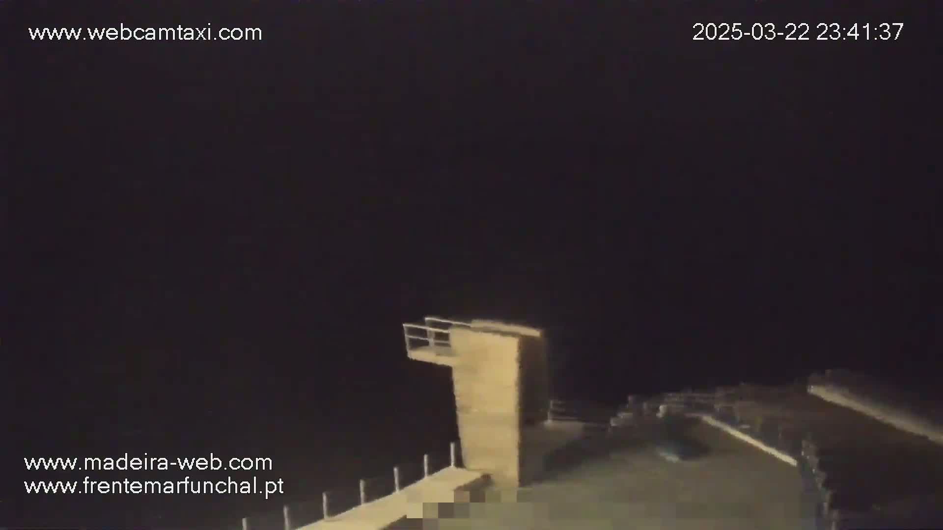 A nighttime, low-light view shows a wooden diving platform extending from a dock overlooking dark water under a completely dark sky.