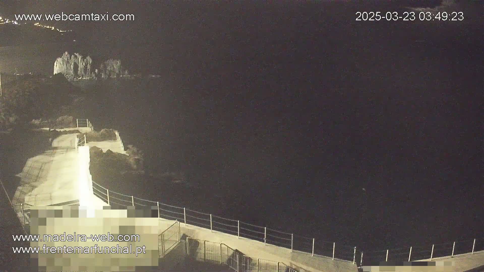 A nighttime, overhead view shows a paved walkway with a railing overlooking a dark body of water, with a rocky outcrop visible in the distance under a clear, dark sky.