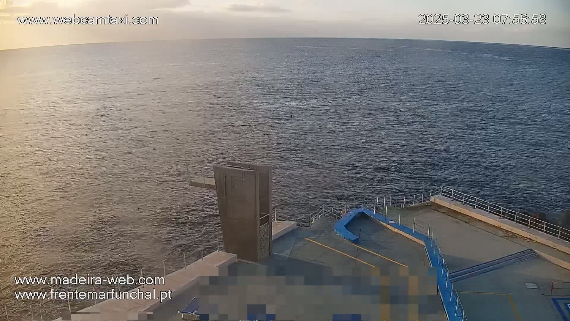 A calm ocean is viewed from an elevated vantage point, showing a diving platform and surrounding concrete area under a partly cloudy sky.