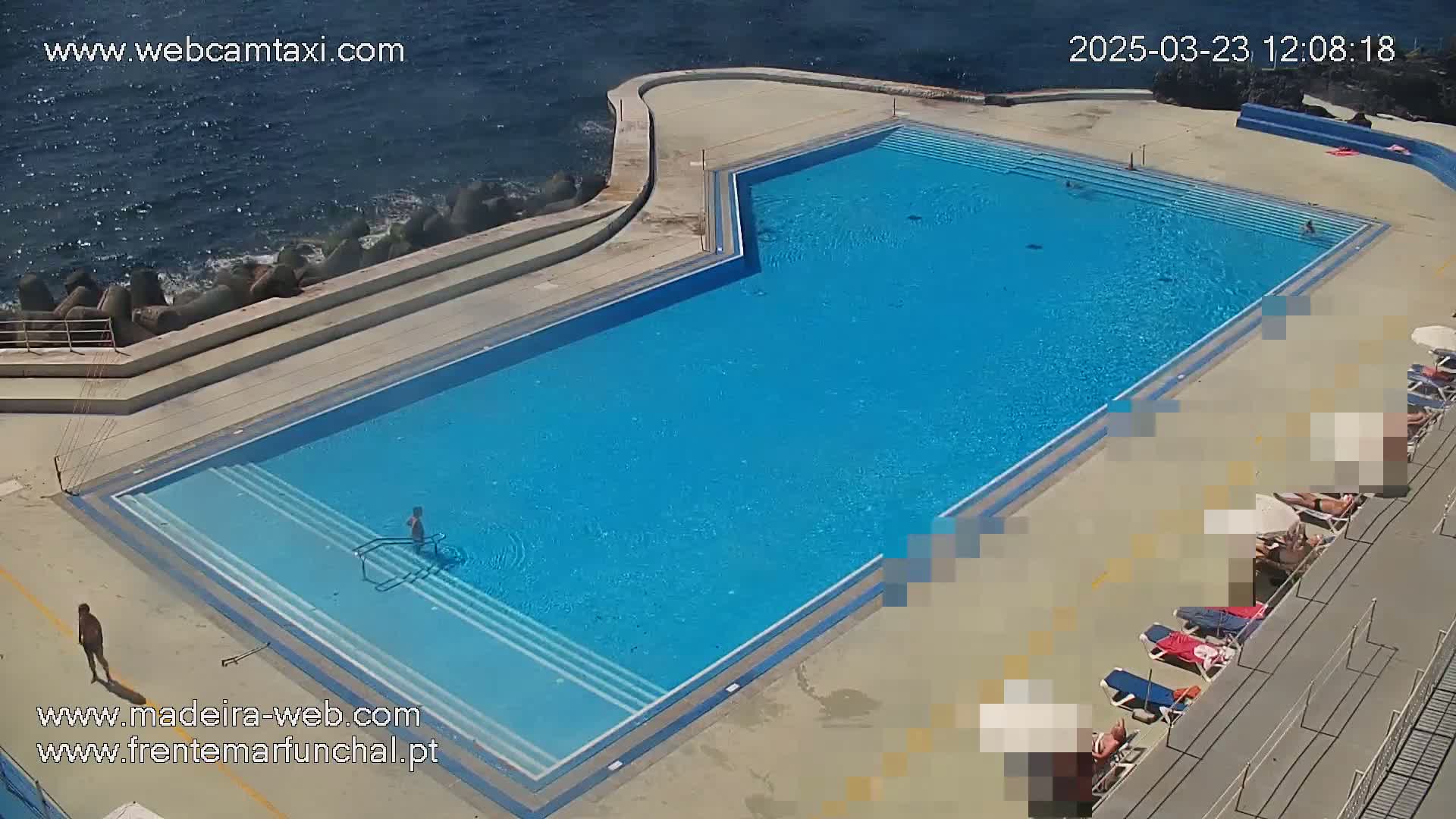 An outdoor pool with several people swimming and sunbathing on a sunny day next to the ocean.