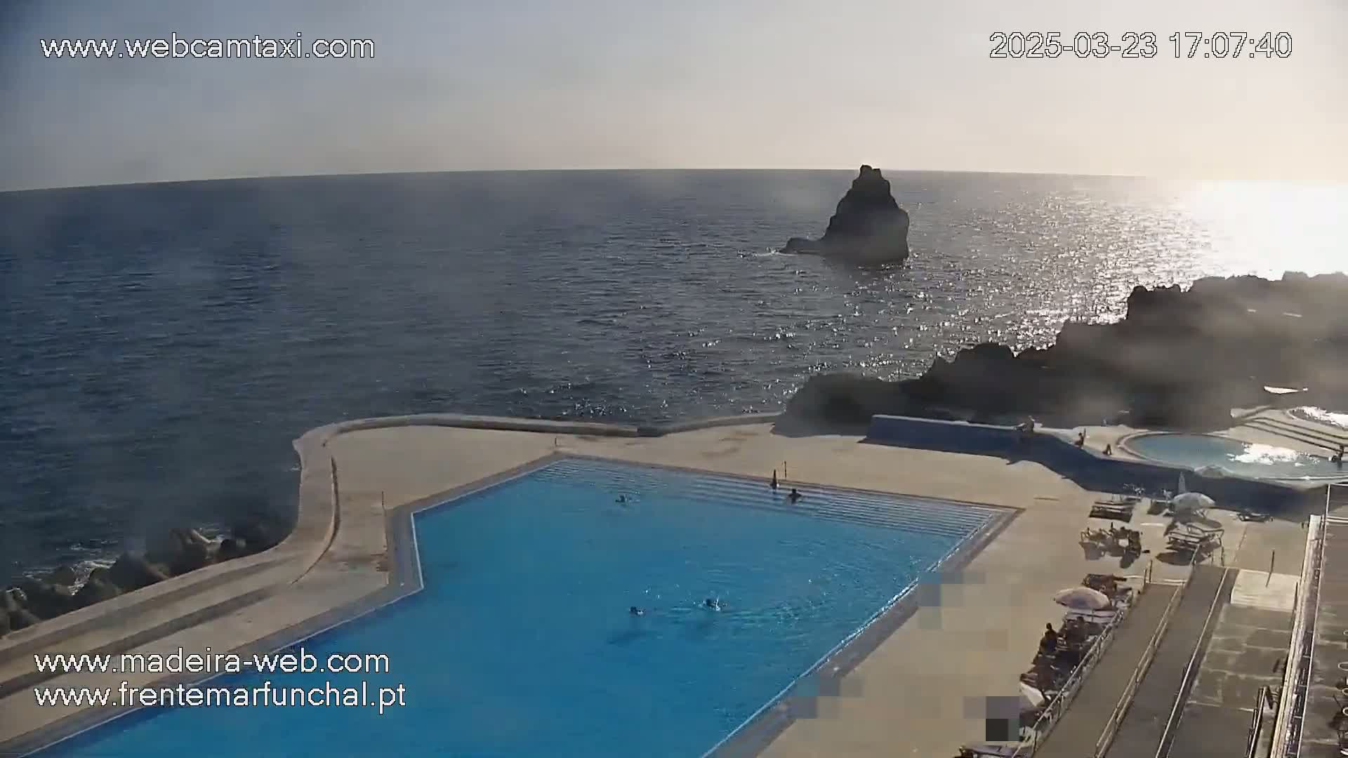 An outdoor pool with several people swimming in it is situated on a rocky coast overlooking a calm ocean under a bright, sunny sky.