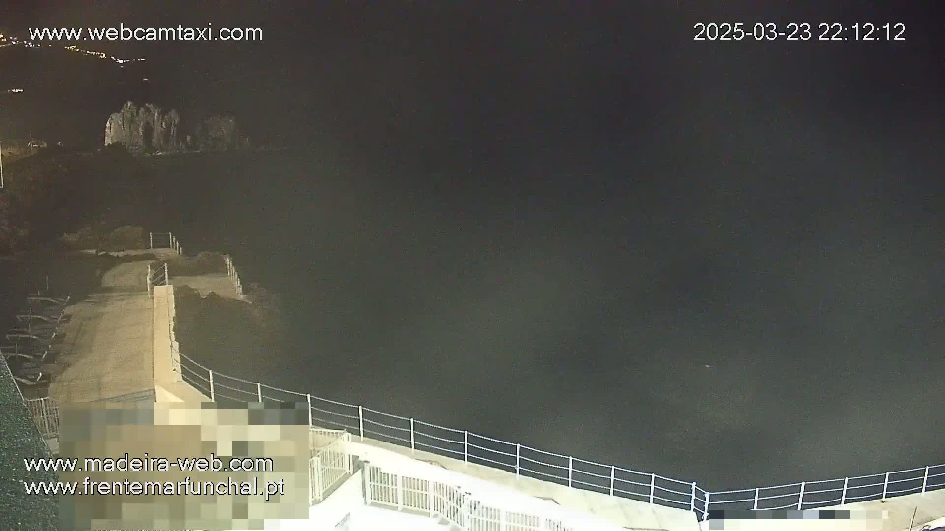 A nighttime view of a concrete walkway and railing overlooking dark water, with a rocky shoreline visible in the distance under a clear, dark sky.