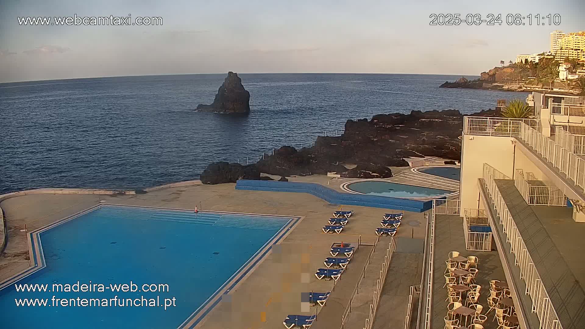 An outdoor scene shows a calm ocean with dark volcanic rock formations, next to a building with multiple pools and lounge chairs, under a clear sky.