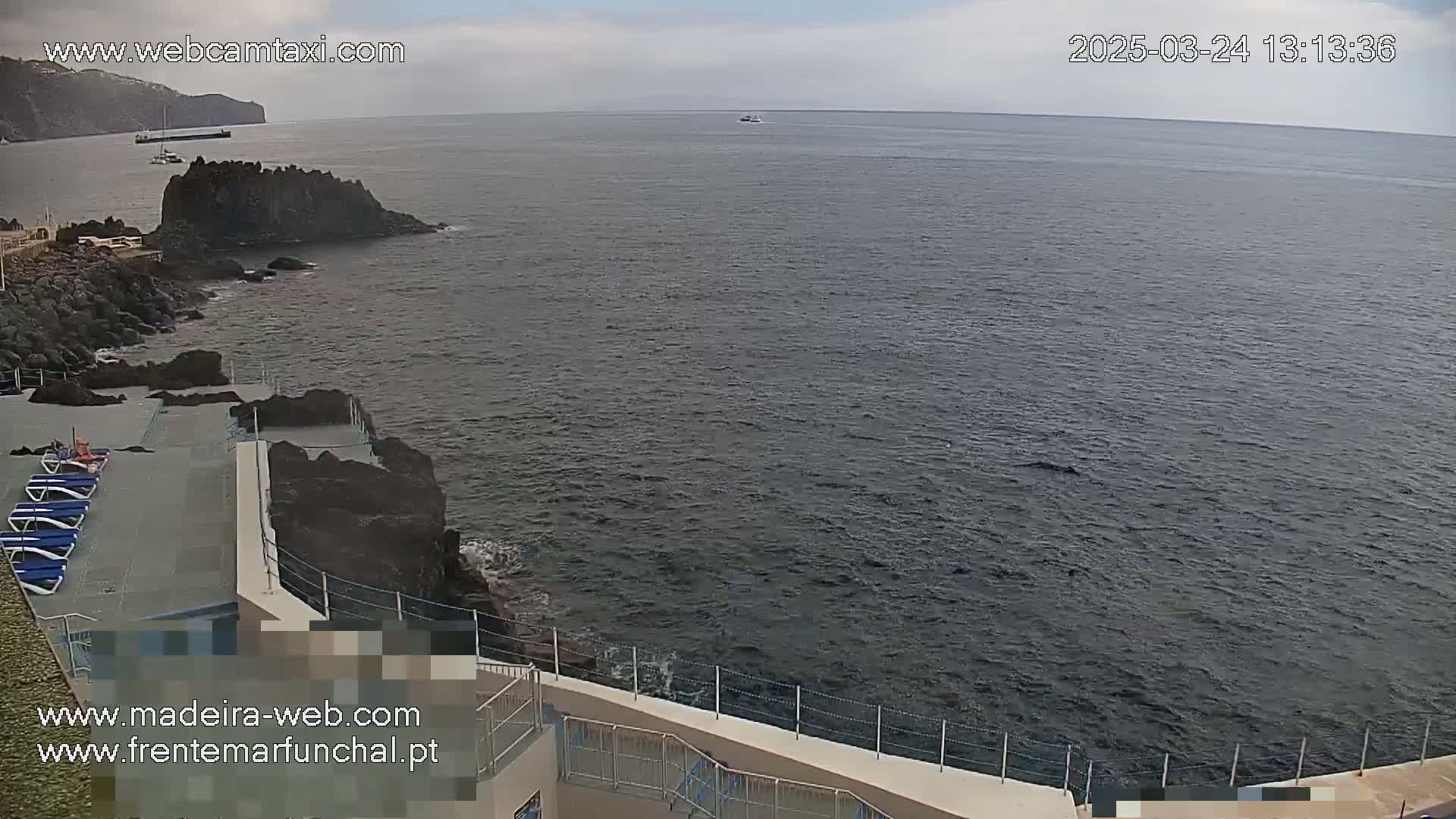 A mostly sunny day shows a rocky coast with a small concrete platform holding lounge chairs overlooking a dark-blue ocean, where a few small boats are visible in the distance.