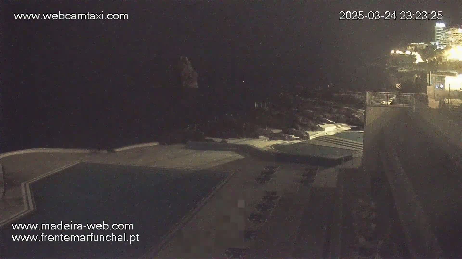 A nighttime, aerial view shows a dark swimming pool, a parking lot, and a building overlooking a dark ocean under a mostly clear sky.