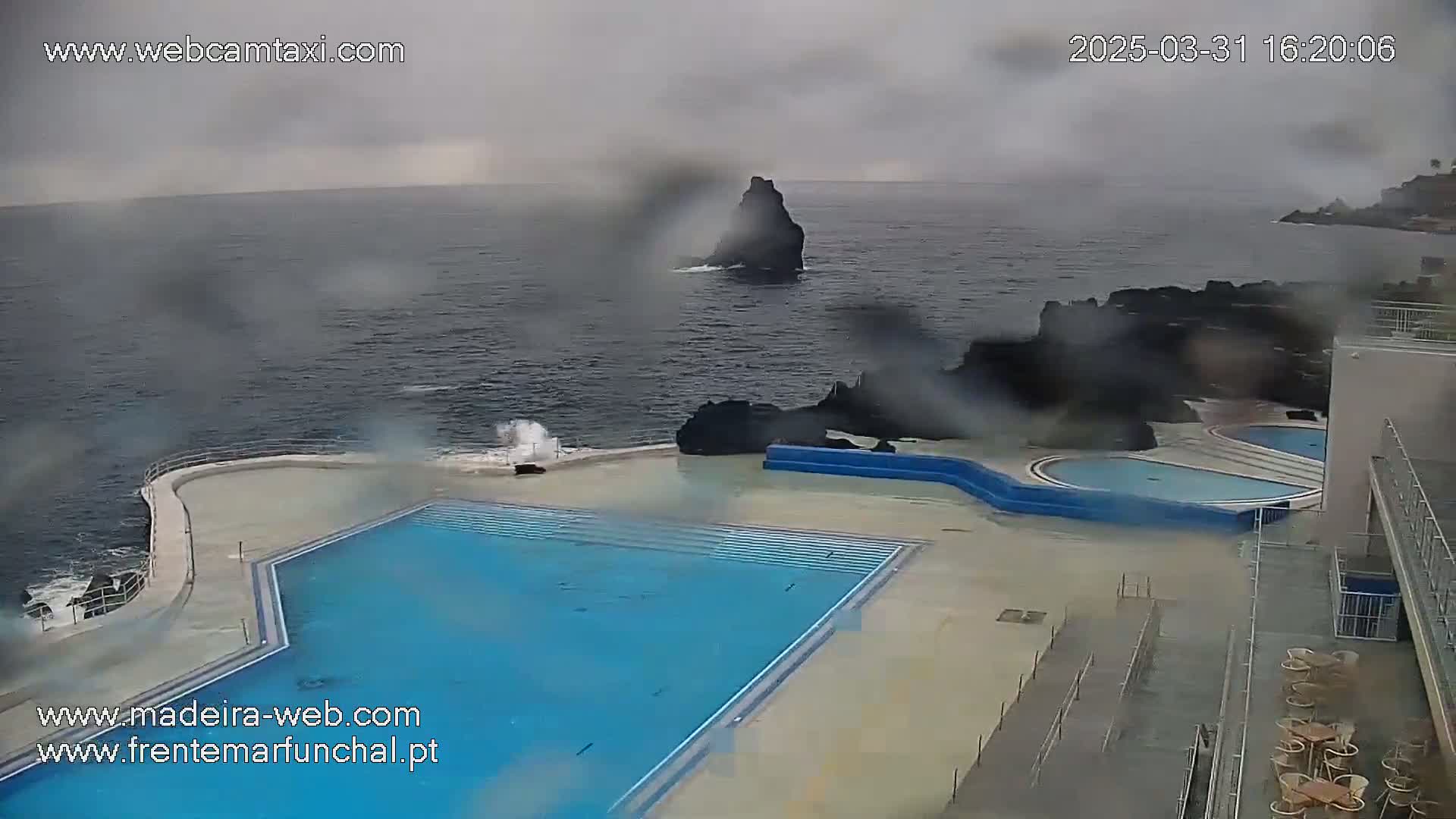 A large outdoor pool sits beside a rocky ocean coastline in rainy weather; waves crash against the rocks near the pool.