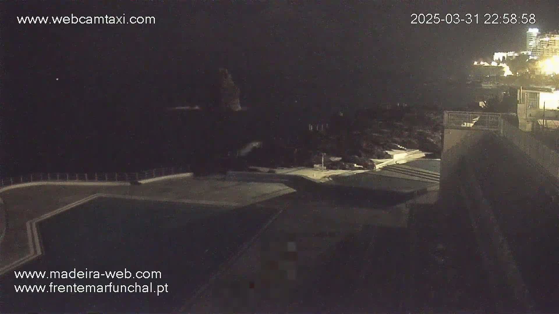 A nighttime, high-angle view shows a dark pool and surrounding buildings next to a dimly lit coastline.