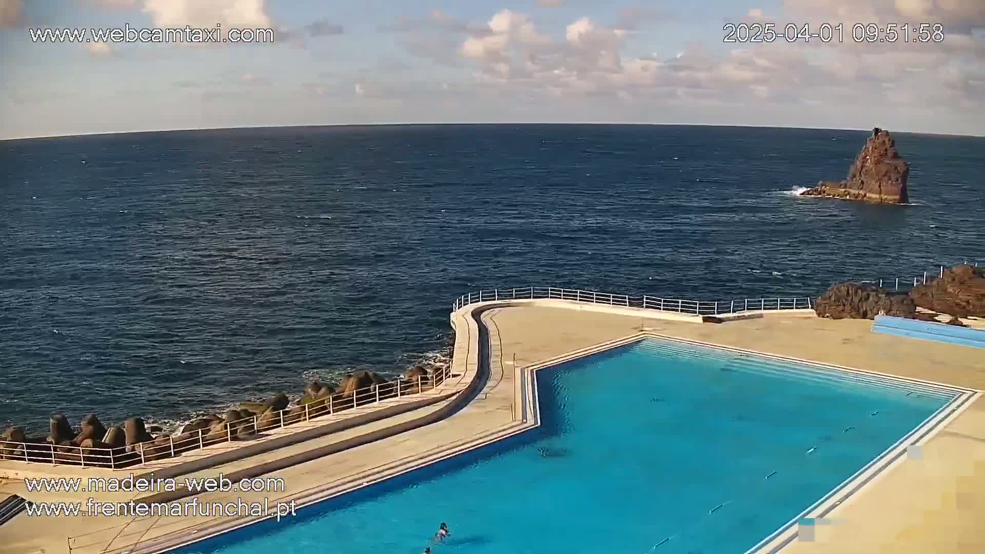 An outdoor pool with light blue water is situated on a rocky coast overlooking a calm ocean under a partly cloudy sky.