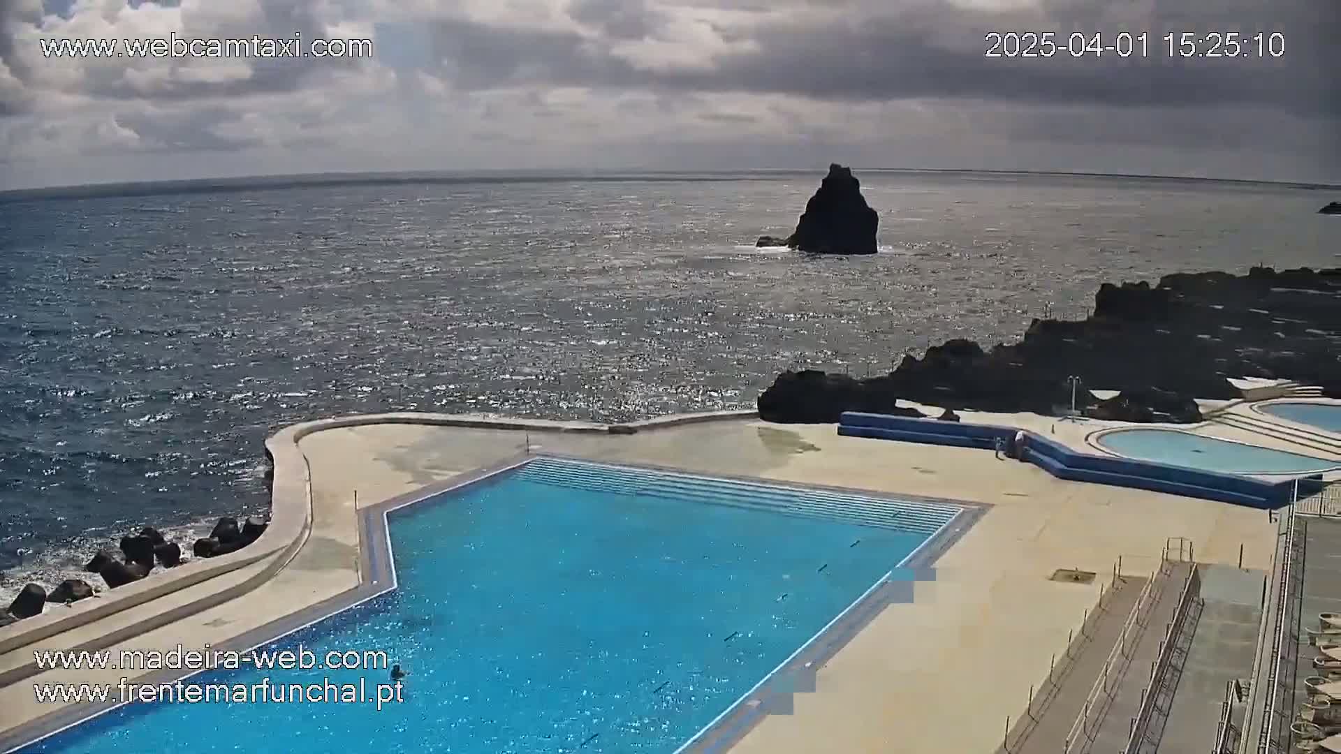 A large outdoor pool complex sits on a rocky coast overlooking a wavy ocean under a partly cloudy sky.