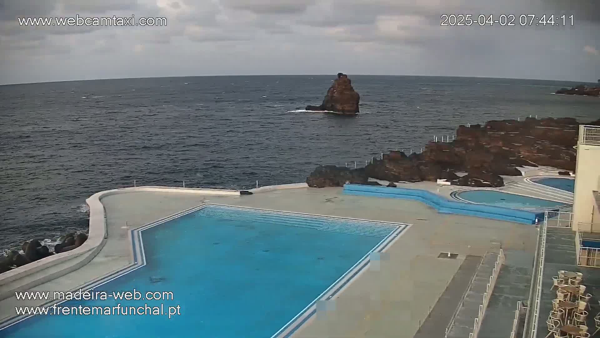 An outdoor pool complex with a large main pool and several smaller pools sits on a rocky coast overlooking a dark, somewhat choppy ocean under a cloudy sky.